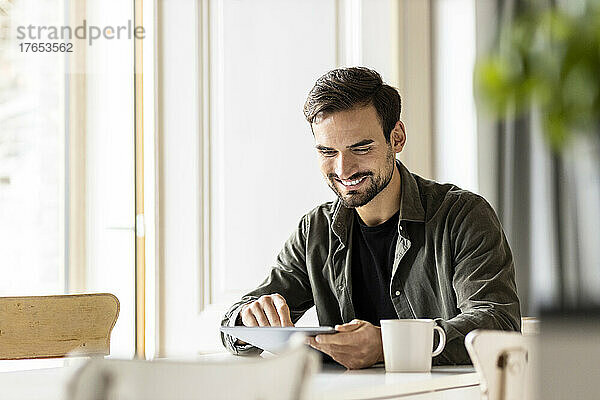 Lächelnder Mann benutzt Tablet mit Tasse auf dem Tisch zu Hause