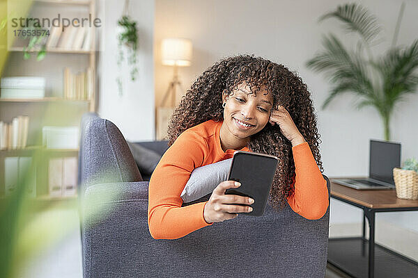 Lächelnde Frau mit lockigem Haar  die zu Hause auf dem Sofa ihr Mobiltelefon benutzt