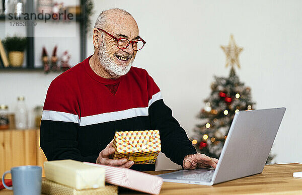 Glücklicher älterer Mann mit Brille und Geschenkbox mit Laptop zu Hause