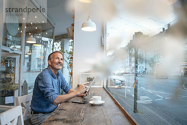 Glücklicher Geschäftsmann mit Tablet-PC  der am Tisch vor der Glasscheibe im Café sitzt