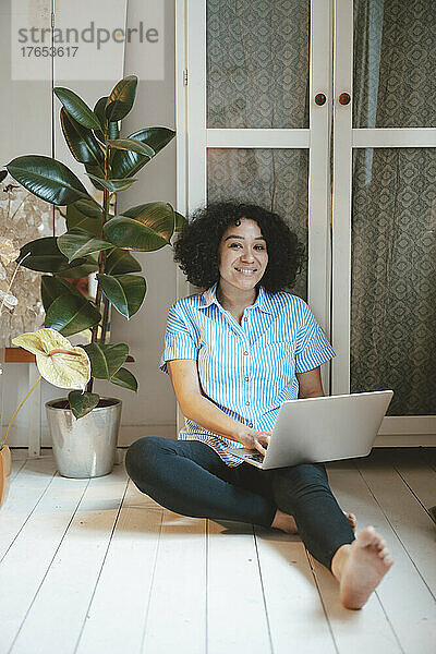 Lächelnde Frau mit Laptop sitzt zu Hause auf dem Dielenbrett