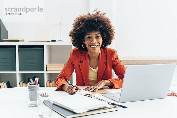 Lächelnde Geschäftsfrau mit Afro-Frisur sitzt mit Laptop am Schreibtisch im Büro