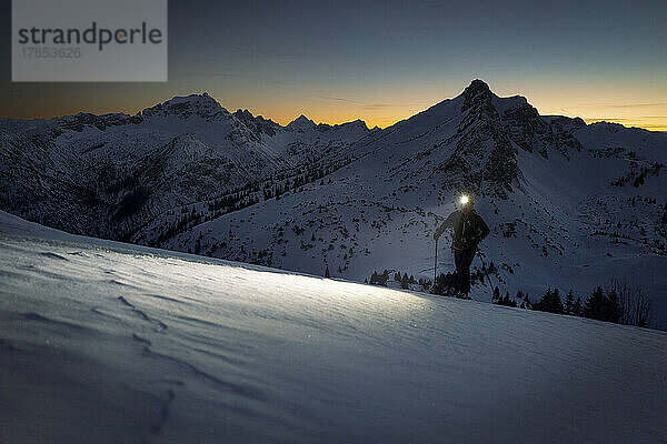 Mann mit Stirnlampe geht bei Sonnenuntergang auf verschneiten Berg