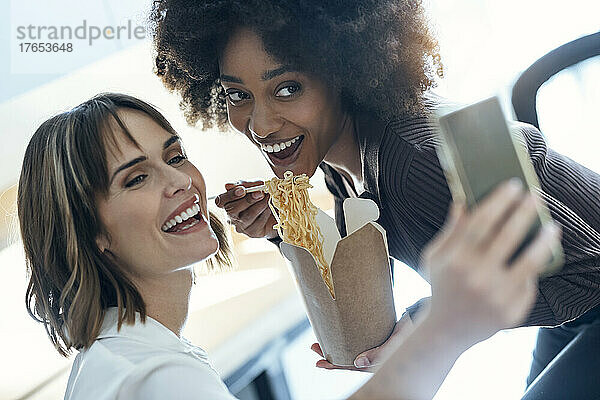 Glückliche Geschäftsfrau macht ein Selfie mit einem Kollegen  der im Büro Nudeln isst