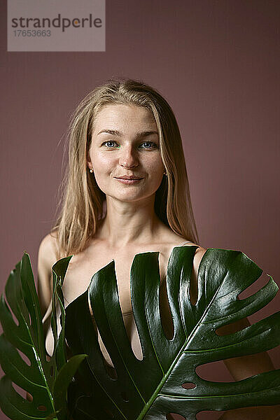 Smiling blond woman with monstera leaves standing against colored background
