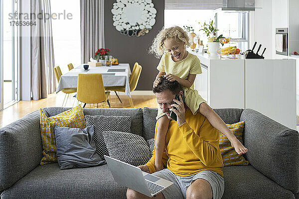 Verspielte Tochter sitzt auf der Schulter des Vaters und telefoniert zu Hause mit dem Laptop