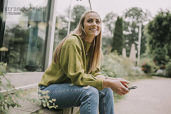 Nachdenkliche junge Frau mit Smartphone sitzt an der Wand