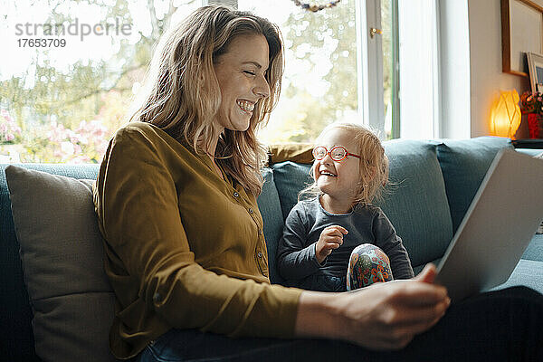 Fröhliche blonde Frau mit Tablet-PC sitzt zu Hause neben ihrer Tochter auf dem Sofa