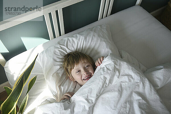 Glücklicher Junge mit Decke  der zu Hause im Bett liegt