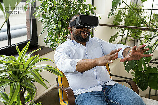 Lächelnder Geschäftsmann mit Virtual-Reality-Simulator sitzt auf einem Stuhl im Büro