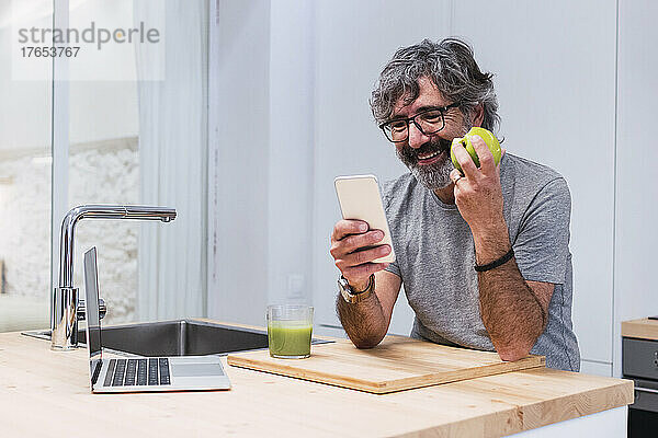 Glücklicher älterer Mann mit Apfel und Smartphone auf der Kücheninsel