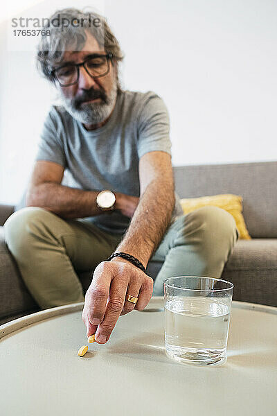 Älterer Mann mit einem Glas Wasser und einer Medikamententablette  die zu Hause auf dem Sofa sitzt