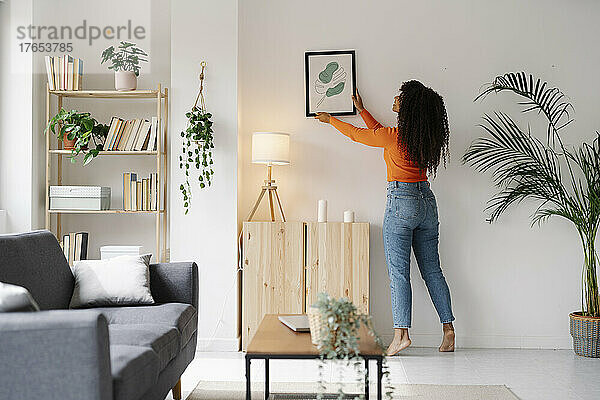 Junge Frau hängt Bilderrahmen an Wand im Wohnzimmer zu Hause
