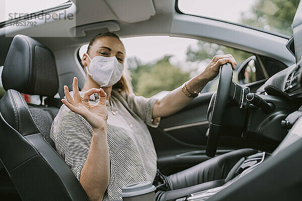 Frau trägt Schutzmaske und zeigt im Auto die Geste „OK“.
