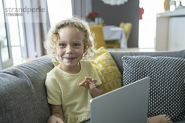 Happy blond girl with laptop sitting on sofa in living room at home