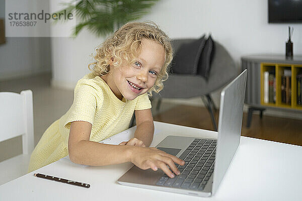 Glückliches blondes Mädchen mit Laptop sitzt am Tisch im Wohnzimmer