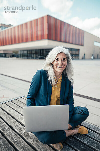 Glückliche Geschäftsfrau mit Laptop sitzt im Schneidersitz im Büropark