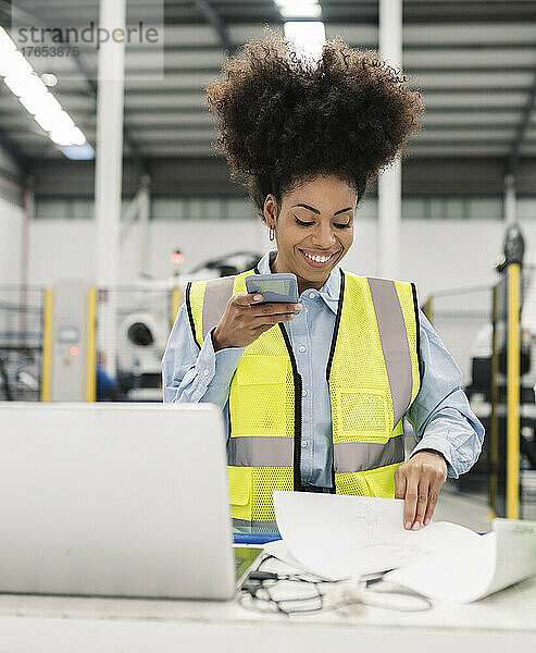 Lächelnder Ingenieur  der Voicemail über sein Smartphone sendet und Dokumente in der Fabrik analysiert