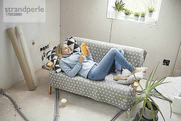 Smiling blond woman lying down on sofa and reading book in attic