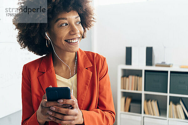 Glückliche Geschäftsfrau hält ihr Mobiltelefon in der Hand und hört im Büro Musik über In-Ear-Kopfhörer