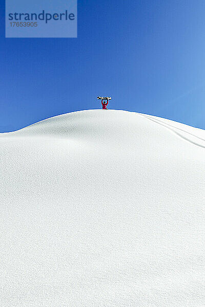 Mann hält Snowboard und steht mit erhobenen Armen am schneebedeckten Berg