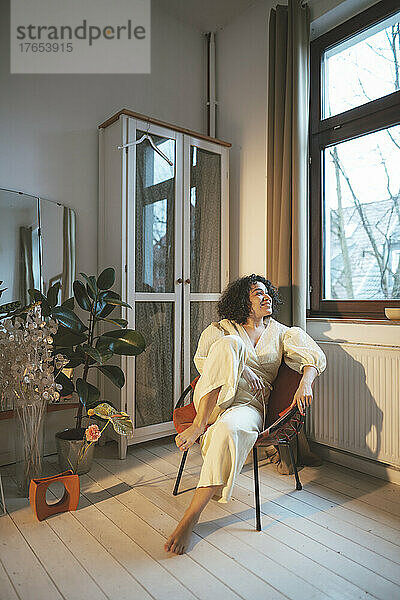 Lächelnde Frau sitzt auf einem Stuhl und schaut zu Hause durch das Fenster