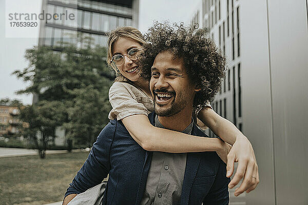 Glücklicher junger Mann  der seine Freundin huckepack vor einem modernen Gebäude trägt