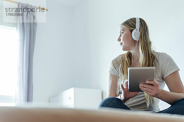 Frau hört Musik über Kopfhörer und sitzt mit Tablet-PC zu Hause im Bett