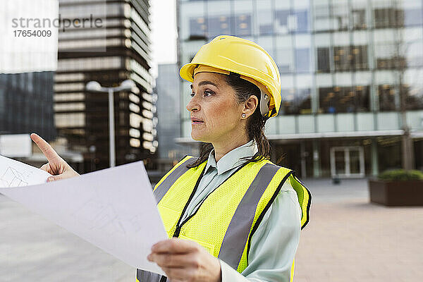 Architekt trägt Schutzhelm und hält einen Bauplan in der Hand  der vor einem modernen Gebäude arbeitet