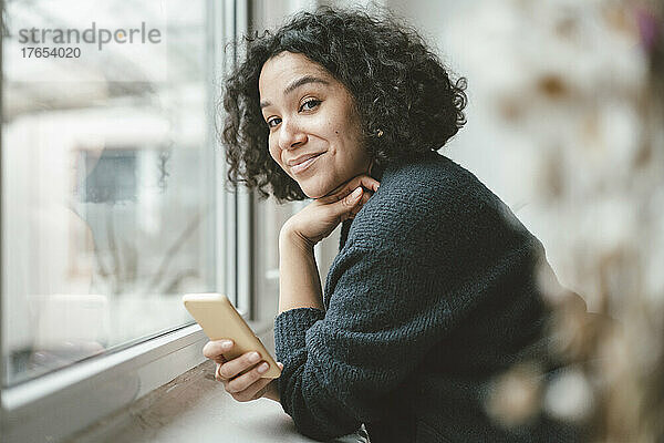Lächelnde schöne Frau lehnt zu Hause mit Mobiltelefon auf der Fensterbank