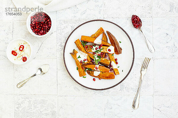 Plate of sweet potatoes with parsley  pomegranate seeds and cumin yogurt lying on white tiles