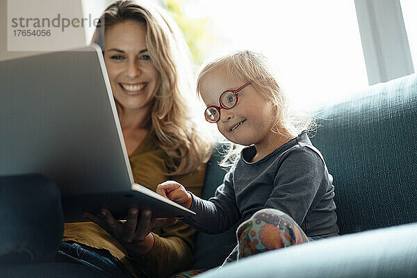 Kleines Mädchen mit Brille und Laptop sitzt neben der Mutter auf dem Sofa im Wohnzimmer