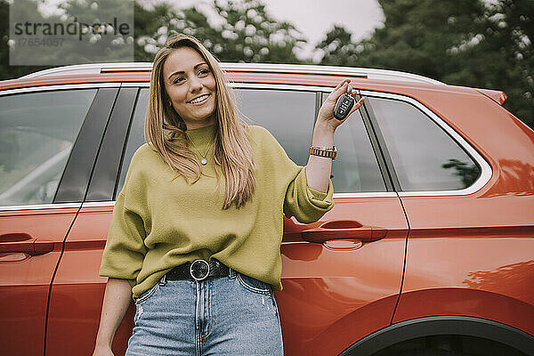 Lächelnde junge Frau mit Schlüssel steht vor dem Auto