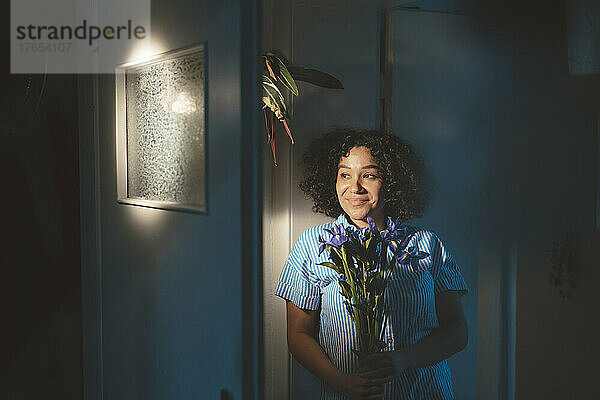 Lächelnde Frau hält Blumen in der Hand und lehnt zu Hause an der Wand