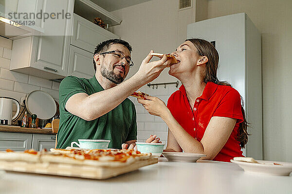 Mann füttert Frau  die am Tisch in der Küche sitzt  mit Pizza