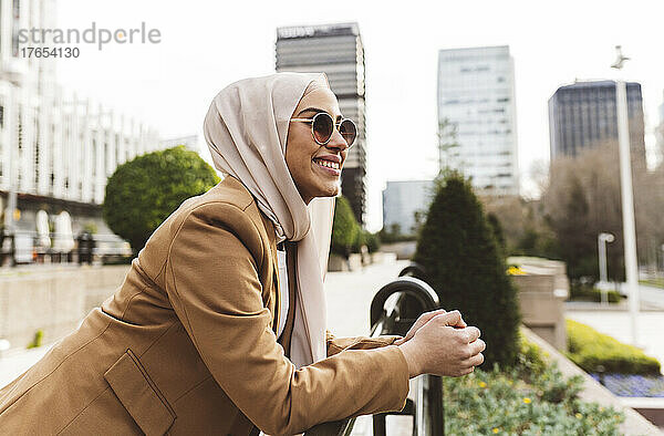 Glückliche Frau mit Sonnenbrille lehnt am Geländer im Büropark