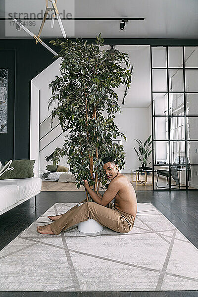 Hemdloser Mann mit geschlossenen Augen umarmt Zimmerpflanze im heimischen Wohnzimmer