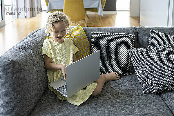 Lächelndes blondes Mädchen mit Laptop sitzt zu Hause auf dem Sofa im Wohnzimmer