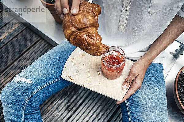 Frau mit Croissant und Marmeladenglas sitzt auf Balkon