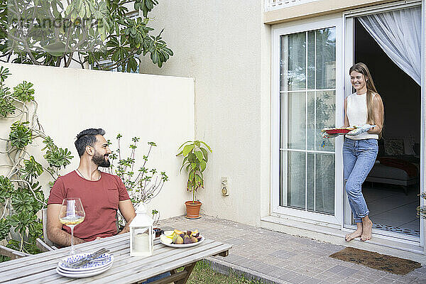 Glückliche Frau trägt herzhaften Kuchen und geht auf Mann zu  der am Tisch im Garten sitzt