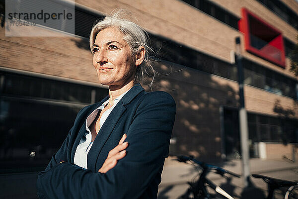Selbstbewusste Geschäftsfrau steht an einem sonnigen Tag mit verschränkten Armen im Büropark