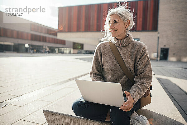 Lächelnde Geschäftsfrau mit Laptop sitzt auf einem Sitz im Büropark