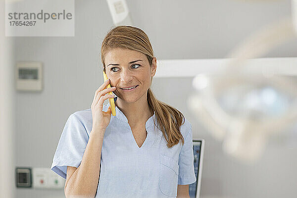 Lächelnder blonder Zahnarzt  der in der medizinischen Klinik mit dem Mobiltelefon spricht