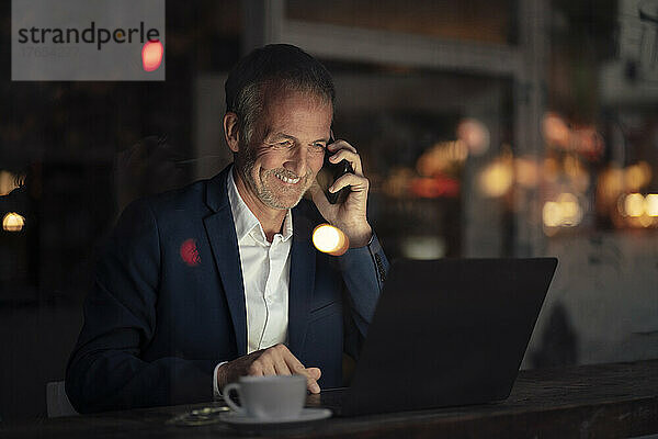 Happy businessman talking on mobile phone and using laptop at night