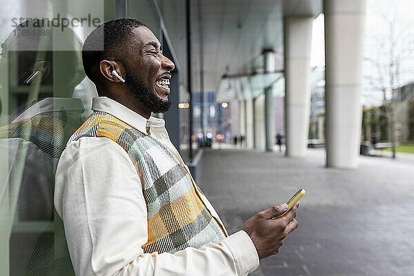 Glücklicher Mann mit geschlossenen Augen  der sein Mobiltelefon an eine Glaswand lehnt