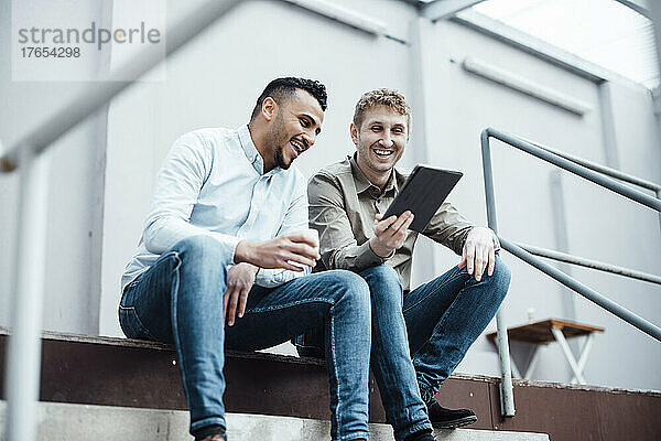 Happy businessman showing tablet PC to colleague siting by railing at work place