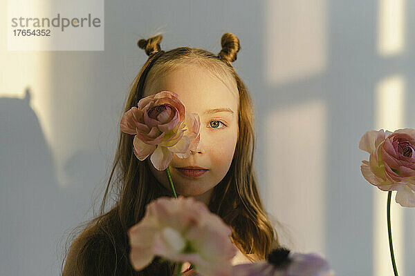 Nettes Mädchen mit Blume im Gesicht  das an einer weißen Wand steht