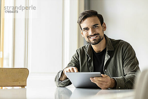 Lächelnder Mann sitzt zu Hause mit Tablet-PC am Tisch