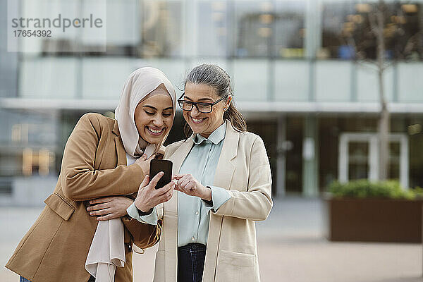 Glückliche Geschäftsfrau mit Brille teilt Smartphone mit Kollege im Büropark