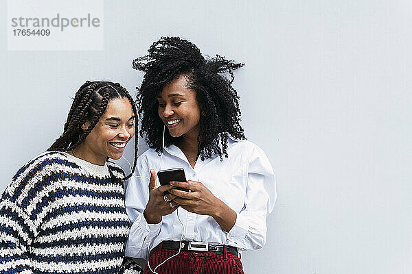 Glückliche Geschäftsfrauen teilen ihr Smartphone und hören Musik über In-Ear-Kopfhörer vor der Wand